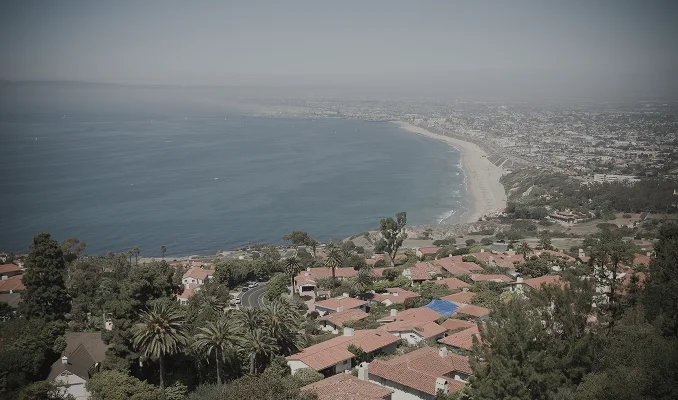 A scenic, high-angle landscape view of a coastal California neighborhood, likely near a beach location. Red-tiled rooftops and lush greenery are in the foreground, with the Pacific Ocean and a hazy coastline stretching into the distance.