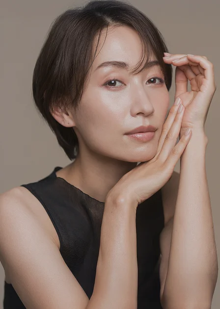A close-up portrait of a woman with a short, dark pixie cut. She is posing with her hands gently framing her face, showcasing her smooth, clear skin and a natural, dewy complexion.