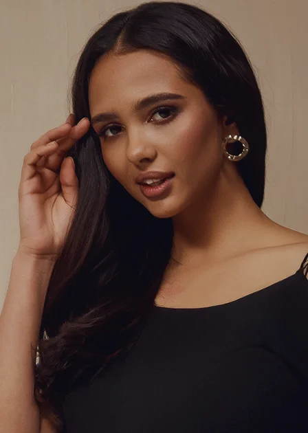A portrait of a woman with long, dark hair and gold hoop earrings. She is looking into the camera with a soft smile and her hand resting near her temple, posing against a warm-toned background.