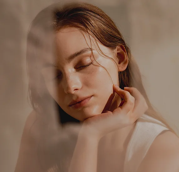 A portrait of a woman with her eyes closed, bathed in warm sunlight with palm leaf shadows across her face, showcasing smooth, healthy skin.