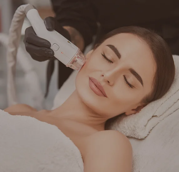 A patient receiving a microneedling or laser skin resurfacing treatment. A practitioner wearing black gloves applies a medical device to the patient's cheek in a bright, clean clinical environment.