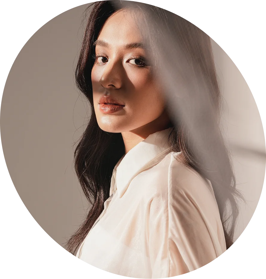 A circular portrait of a young woman with long, dark hair and glowing skin. She is looking into the camera with a soft, confident expression, wearing a white silk blouse.
