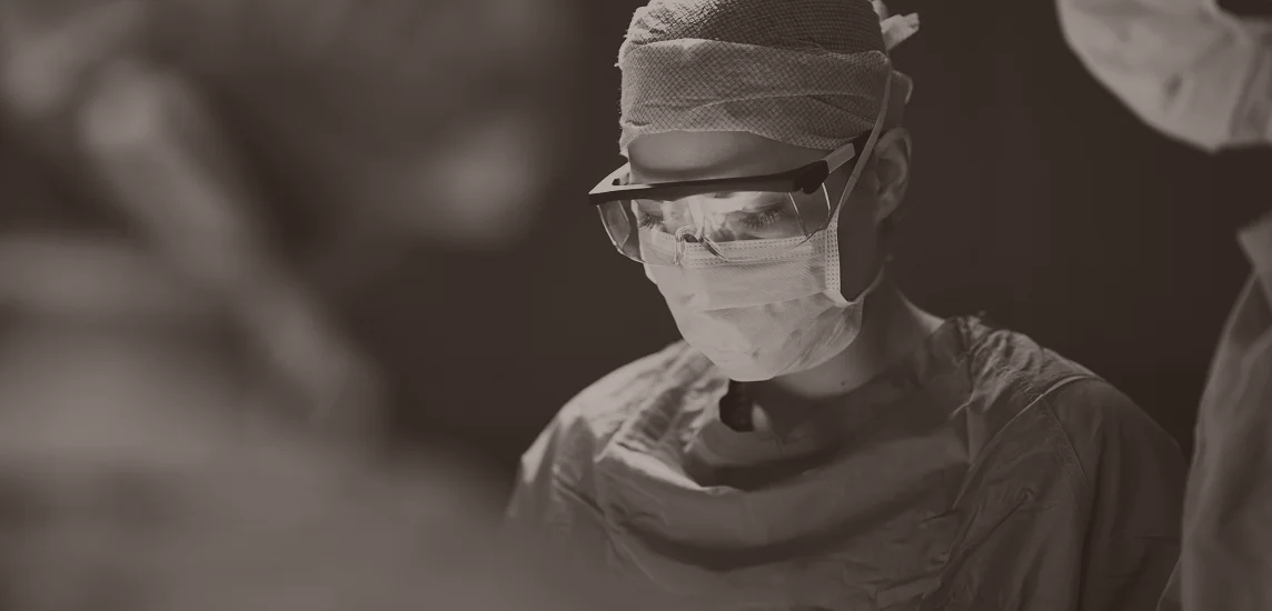 A dramatic, sepia-toned close-up of a surgeon (Dr. Moy) during a reconstructive procedure. She is wearing surgical loupes (magnification glasses), a mask, and a surgical cap, illuminated by a bright overhead light.
