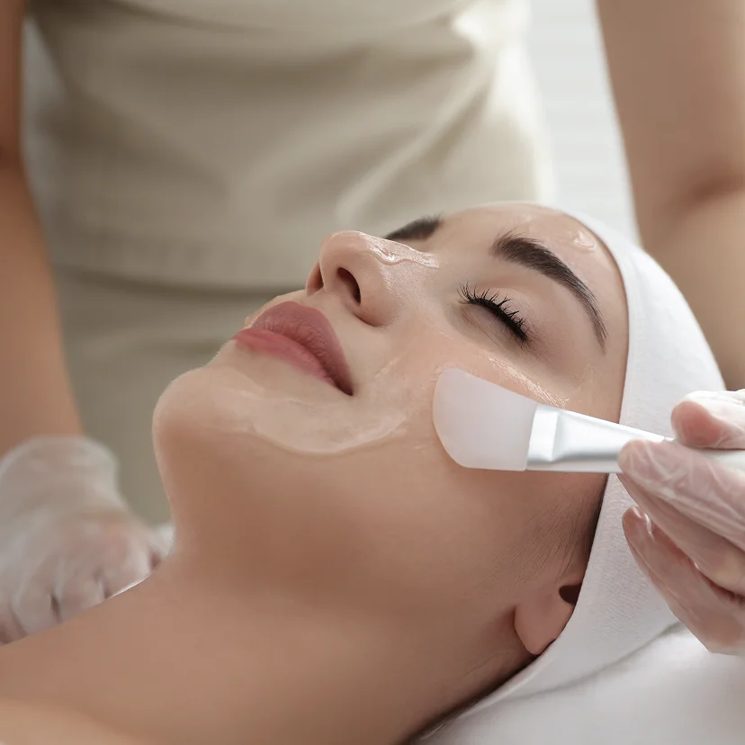 A patient lying down with a white headband receiving a professional facial treatment. A provider is using a soft brush to apply a translucent gel or mask to the patient's cheek.