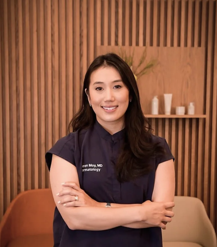 A professional portrait of Dr. Lauren Moy, MD, a dermatologist, smiling with her arms crossed. She is wearing dark blue scrubs in a modern office with light wood-slat wall paneling.
