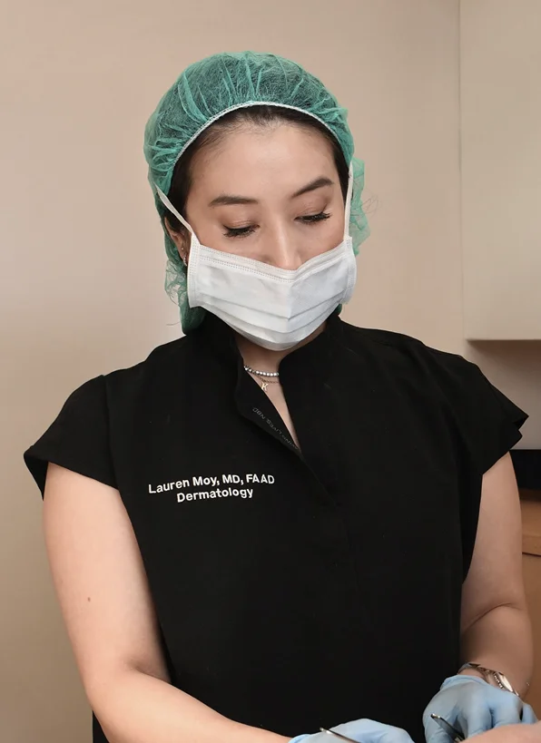 Dr. Lauren Moy, MD, FAAD, wearing black scrubs, a surgical mask, and a green hair net, looking down while performing a precise dermatological procedure.