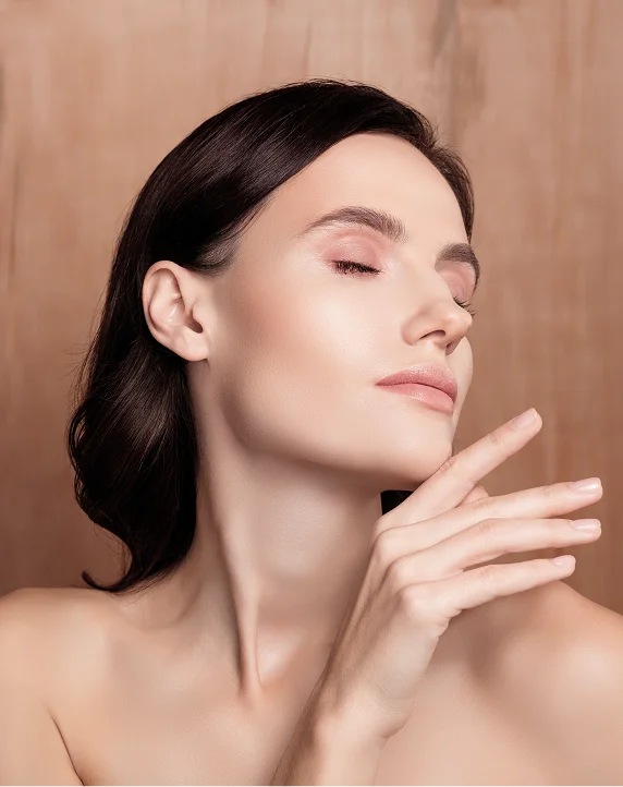 A profile view of a woman with clear, glowing skin and dark hair, posing elegantly with her hand near her chin against a warm, wood-textured background.