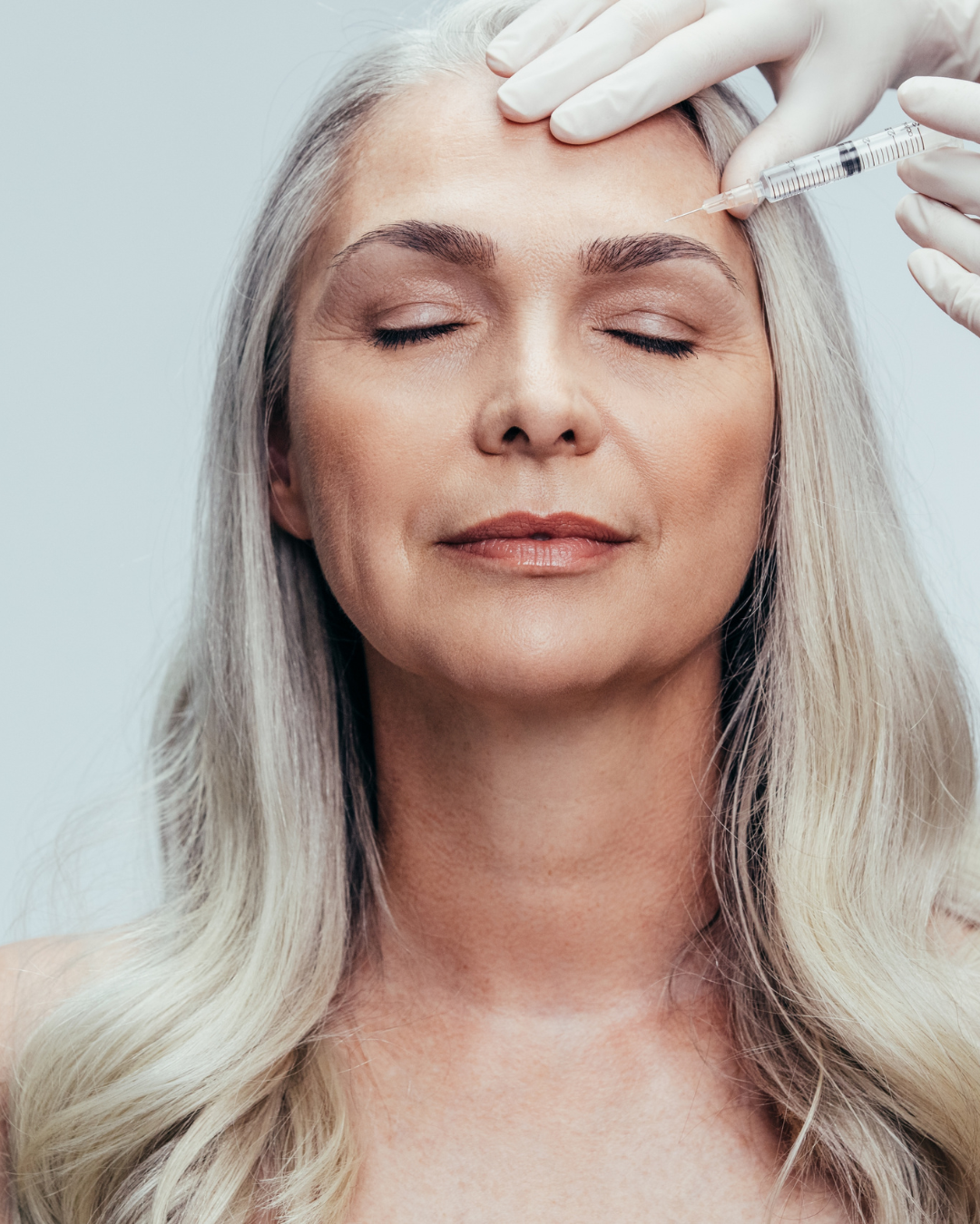 A close-up of a mature woman with grey hair receiving a cosmetic injection near her eyebrow. A practitioner in white gloves (Dr. Moy's team) is carefully administering the treatment.