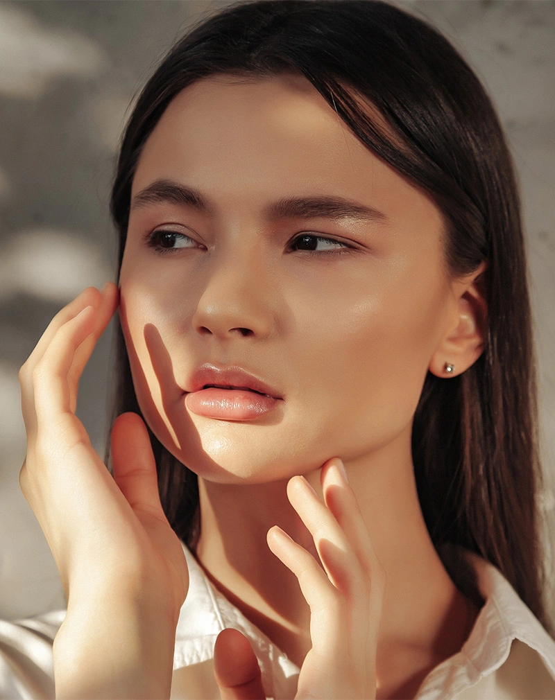 A vertical, close-up portrait of a woman with dark hair and a smooth, luminous complexion. She is gently framing her face with her hands, looking slightly off-camera in soft, natural lighting. - Bio-Advanced Skincare in California.