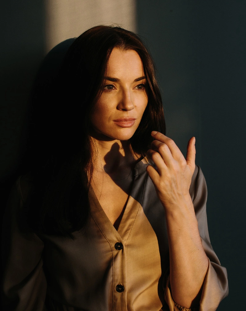 A vertical portrait of a woman with dark hair standing in deep shadow with a strip of golden sunlight illuminating her face and hand. She has a neutral expression and is wearing a button-down blouse. - Botox Wrinkle Relaxers in California.