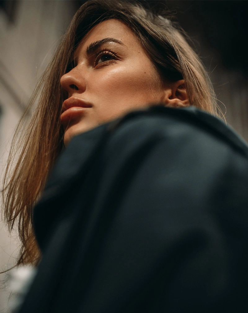 A vertical, low-angle profile shot of a woman looking upward. Her hair is swept back, and the lighting highlights her defined jawline and clear skin against a moody, blurred background. - Brow Lift in California.