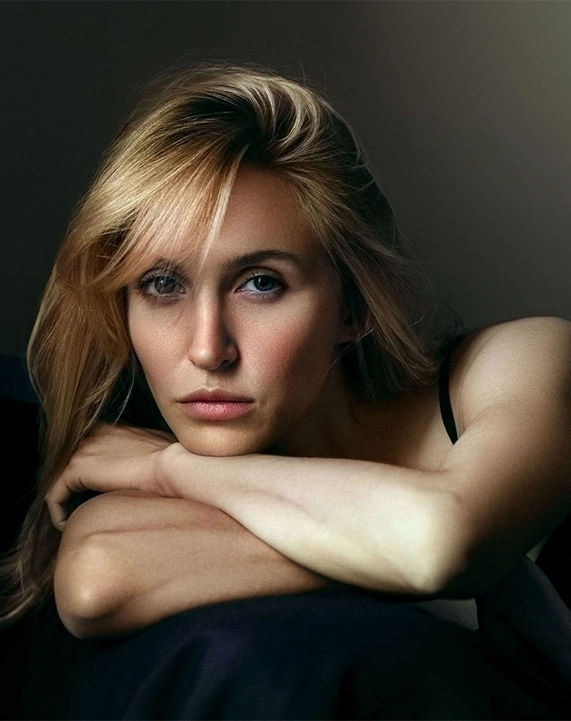 A vertical close-up of a woman with blonde hair resting her chin on her crossed arms. She is looking directly into the camera with a serious expression and striking blue eyes. - Co2 Laser in California.