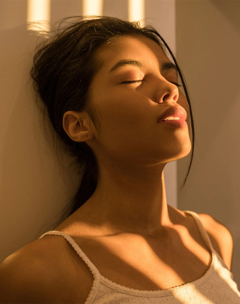 A vertical image of a woman with her eyes closed and head tilted back, leaning against a wall. Warm sunlight dapples across her face and white tank top, creating a peaceful, glowing effect. - Chemical Peels in California.