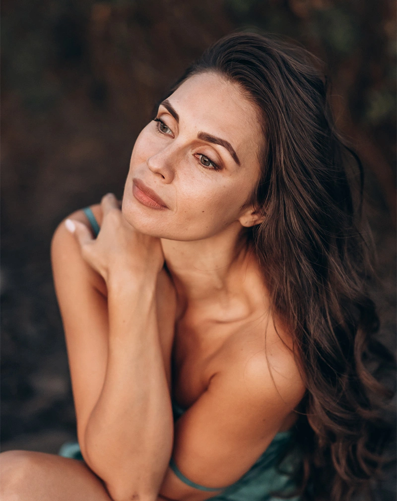 A vertical shot of a woman with long, wavy brown hair looking off into the distance. She is holding her hands to her shoulders in a soft, outdoor setting with warm, diffused light. - Deep Plane Facelift in California.