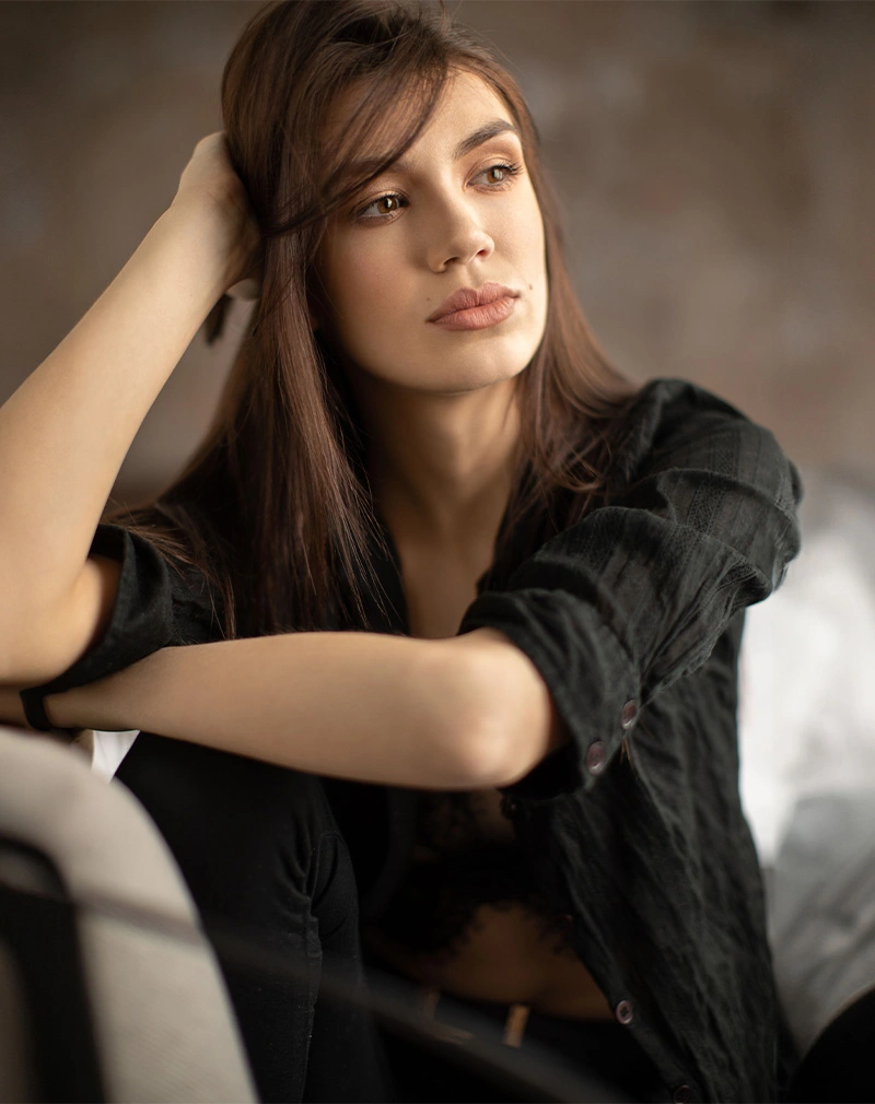 A vertical portrait of a woman with long brown hair wearing a black button-up shirt over a lace top. She is leaning forward with one hand in her hair, looking thoughtfully to the side. - Dermal Fillers in California.