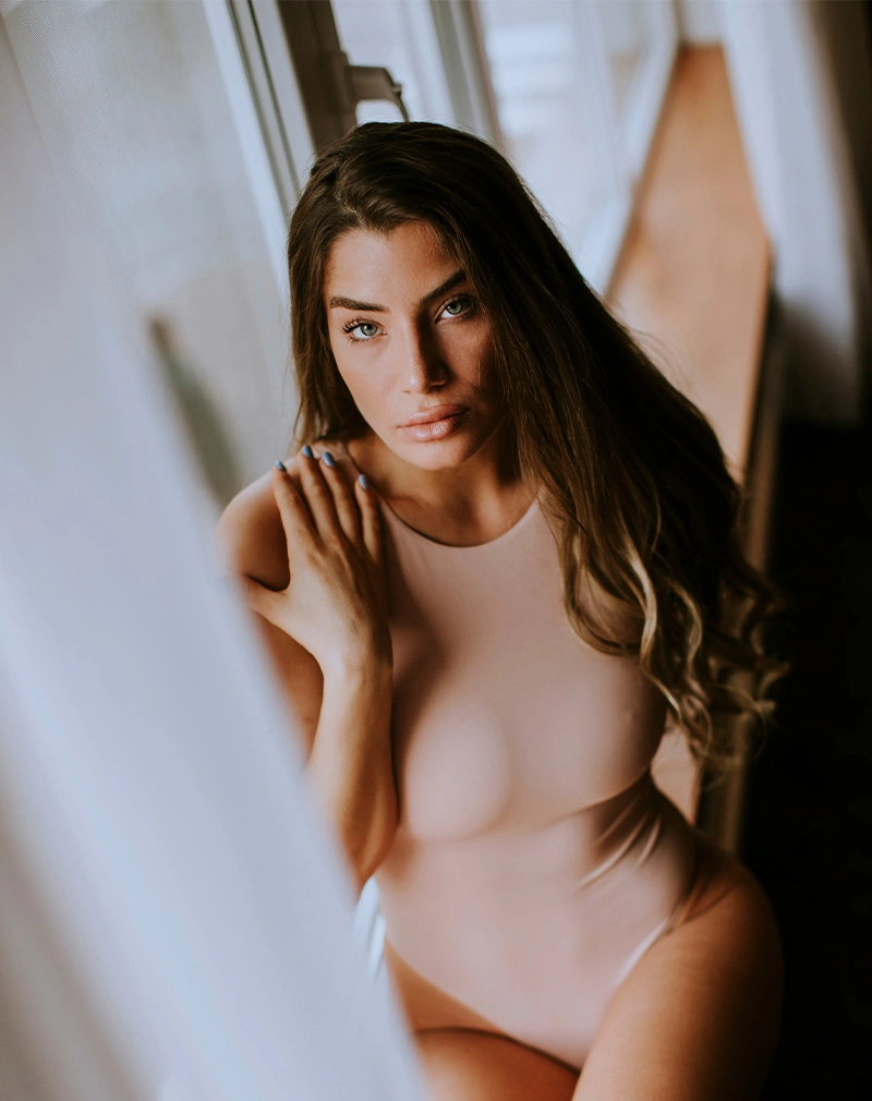 A vertical high-angle shot of a woman with long wavy hair and blue eyes, wearing a nude-colored bodysuit. She is looking directly at the camera with soft, natural lighting coming from a nearby window. - Laser Hair Removal in California.