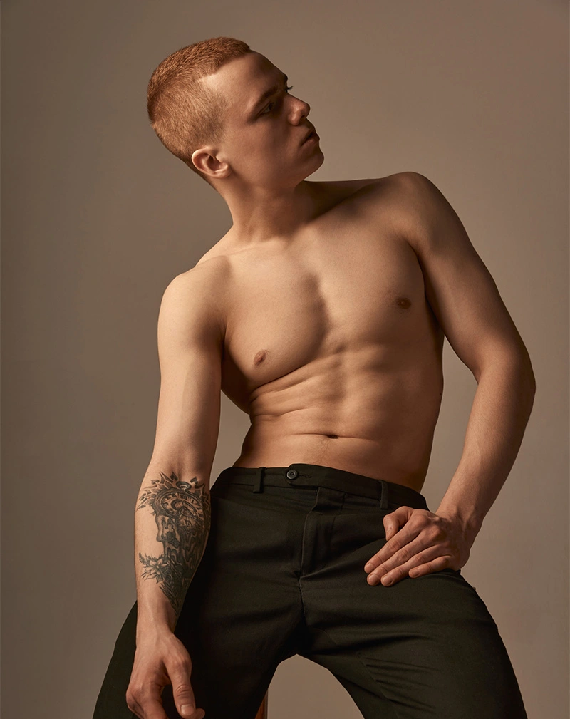 A vertical portrait of a shirtless man with short red hair and a visible tattoo on his forearm. He is wearing black trousers and looking off to the side in a posed studio shot. - Laser Tattoo Removal in California.