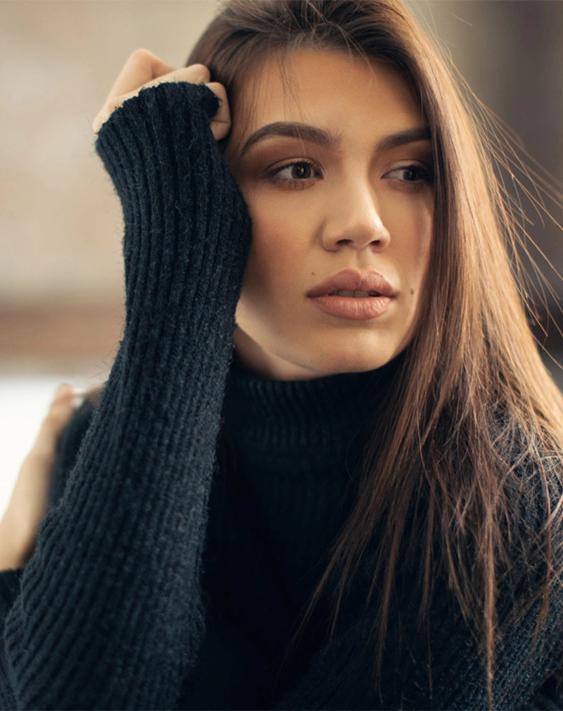 A vertical close-up of a brunette woman wearing a black ribbed turtleneck. She is looking off-camera with a thoughtful expression, with one hand resting near her forehead. - Lower Eyelid Surgery in California.