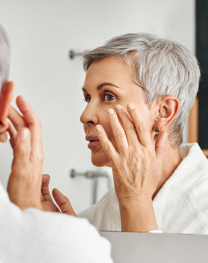 A vertical shot of a woman with short silver hair wearing a white bathrobe, looking into a bathroom mirror while gently touching the skin under her eye. - MOHS Reconstructive Surgery in California.