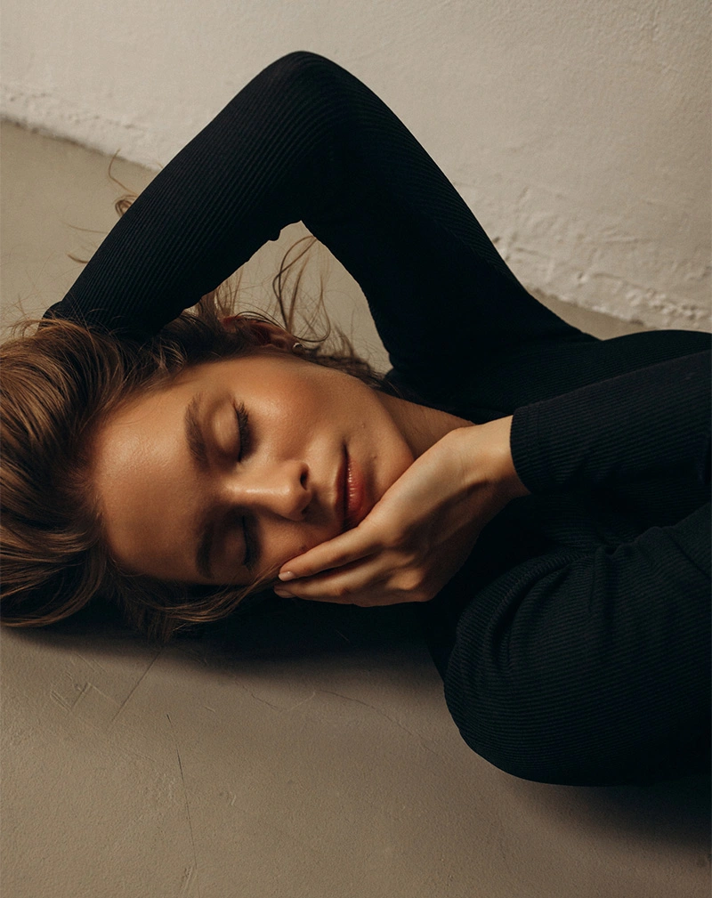 A vertical, moody portrait of a woman lying down with her eyes closed, resting her face against her hand. She is wearing a black ribbed long-sleeve top. - Nano Fat Transfer in California.