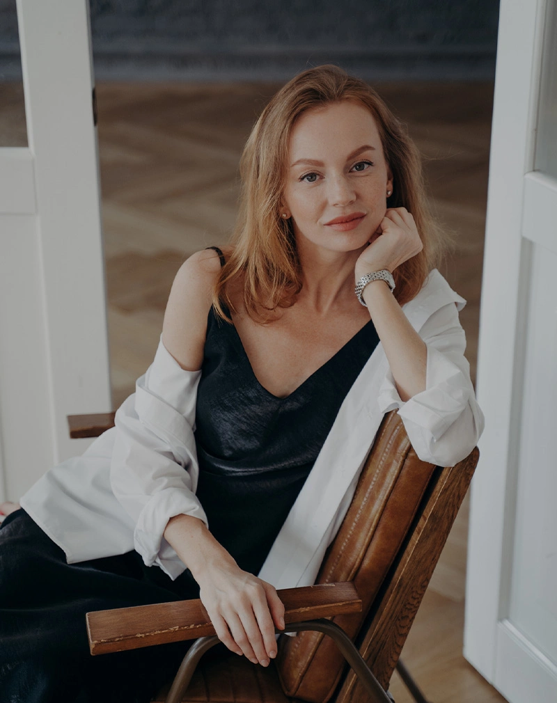 A vertical portrait of a woman with auburn hair and freckles sitting in a leather chair. She is wearing a black silk camisole and a white unbuttoned shirt, resting her chin on her hand. - Neck Lift in California.