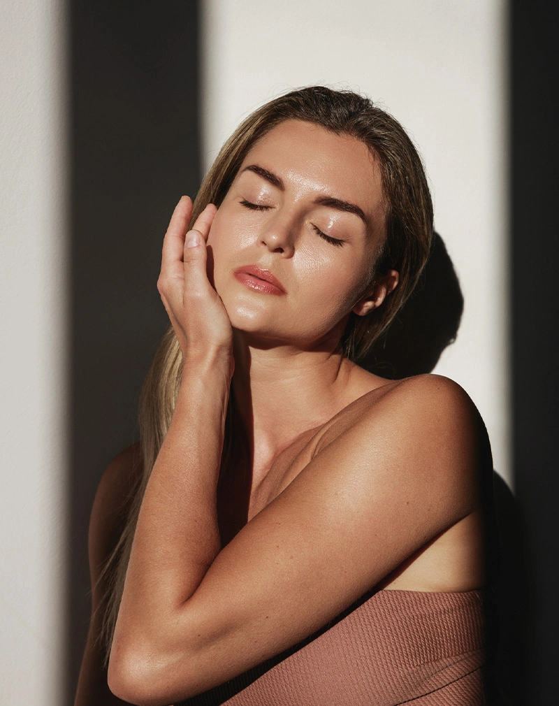 A vertical portrait of a blonde woman with her eyes closed, basking in bright sunlight that creates a strong shadow across the wall behind her. She is touching her cheek with a peaceful expression. - PRP EZ Gel in California.