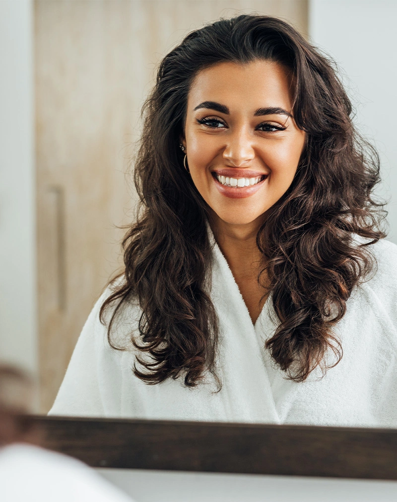 A vertical, bright portrait of a woman with long, voluminous dark curly hair wearing a white bathrobe and smiling widely, showing her teeth. - Pelleve in California.