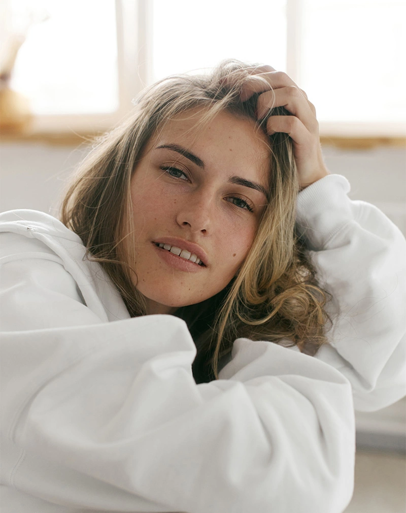 A vertical, candid-style photo of a woman with light brown hair and freckles wearing a white oversized hoodie, looking at the camera with a soft smile while running a hand through her hair. - Pico Laser in California.