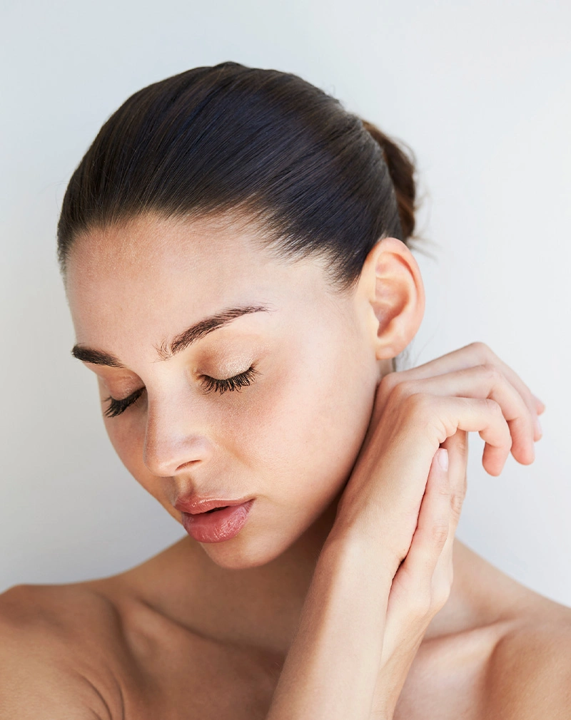 A vertical, close-up portrait of a woman with her hair pulled back and eyes closed, looking downward. Her hands are clasped near her neck against a clean white background. - Radiesse in California.