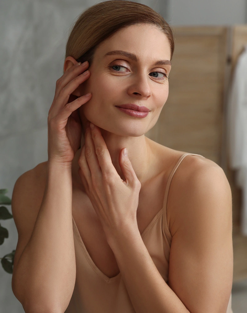 A vertical portrait of a woman with her hair pulled back, smiling softly while touching her cheek and neck area, highlighting her smooth skin. - Surgical Scar Revision in California.