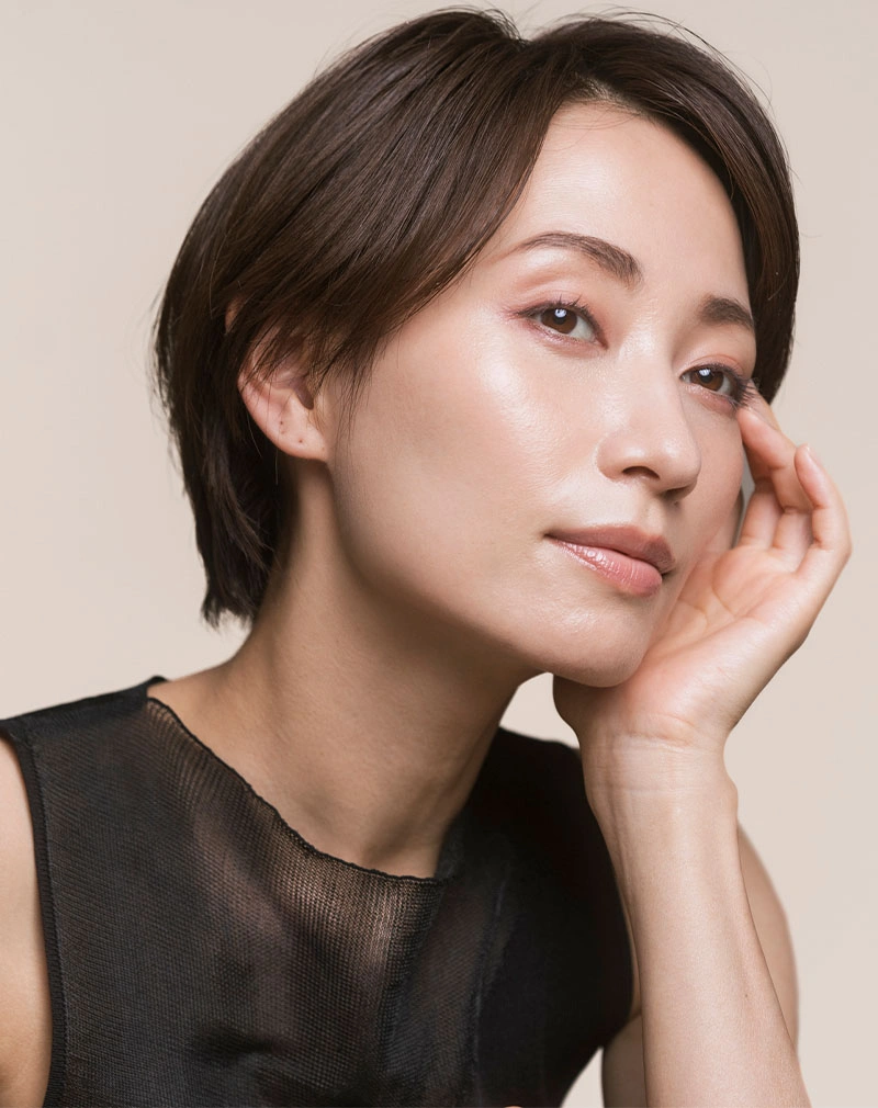 A vertical close-up of a woman with a short dark bob and shimmering eyeshadow, resting her face on her hand and looking slightly off-camera. - Upper Eyelid Surgery in California.
