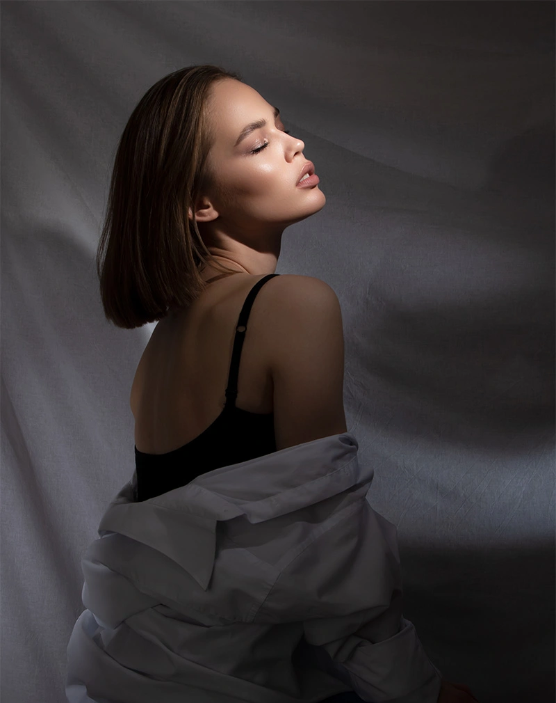 A vertical, moody portrait of a woman with a sleek brown bob, seen from behind with her head turned in profile. She is wearing a black camisole and a white shirt draped off her shoulders, illuminated by a single light source against a draped grey backdrop. - Excessive Sweating in California.