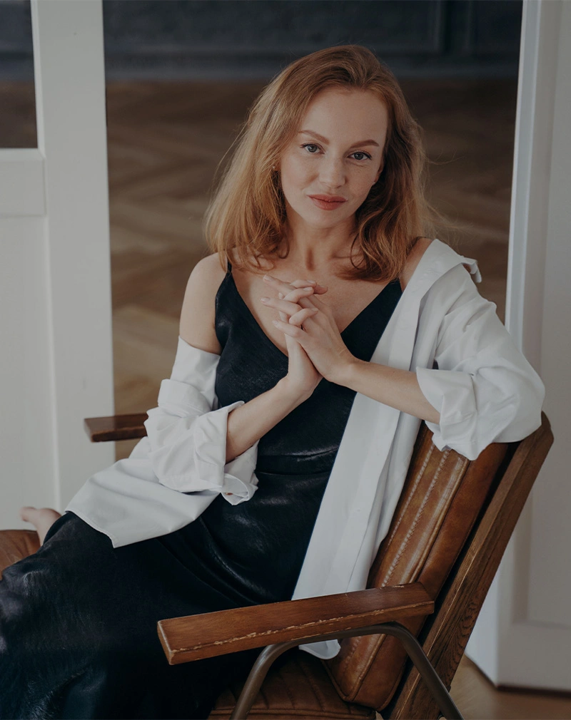A vertical portrait of a woman with auburn hair sitting in a leather chair with her hands clasped. She is wearing a black silk camisole under an unbuttoned white shirt. - Hand Rejuvenation in California.
